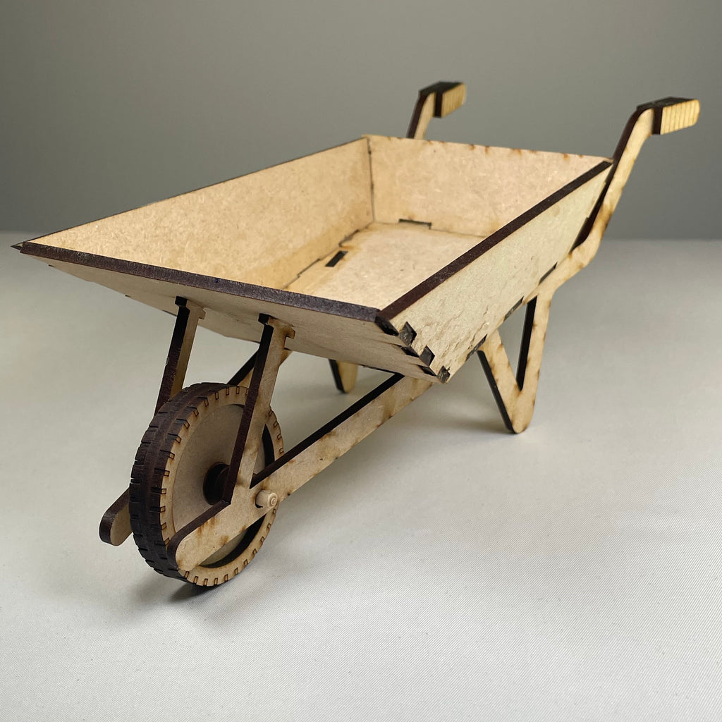 Curious Rabbit Wheelbarrow Planter garden kit, flatpack model with green plants and soil, ready for assembly in a garden setting.