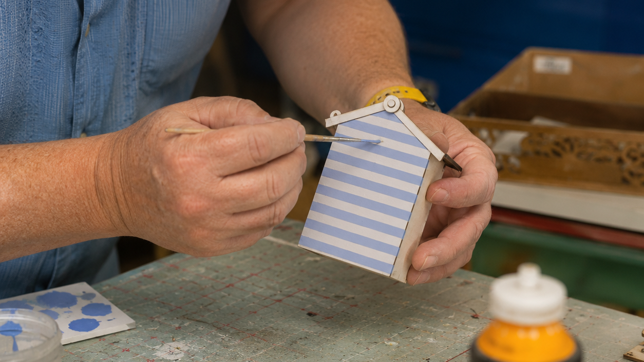 A close-up of a Richard's hands holding a small beach hut with blue and white stripes while applying paint with a brush. Richrad is wearing a blue shirt, and a worktable with paint bottles is visible in the background.